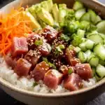 Tuna poke bowl cooked - the image is a close-up of a bowl of food. the bowl is made of ceramic and has a dark brown color. inside the bowl, there is a bed of white rice. on top of the rice, there are several pieces of sliced tuna, sliced avocado, and sliced cucumber. the tuna is covered in sesame seeds and is garnished with chopped cilantro. on the side of the bowl is a pile of shredded carrots. the background is blurred, but it appears to be a black countertop.