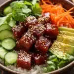Tuna poke bowl cooked - the image is a close-up of a bowl of food. the bowl is made of dark brown ceramic and is placed on a wooden table. the food in the bowl appears to be a stir-fry dish with rice, sliced cucumbers, carrots, and sliced avocado. the tofu is covered in sesame seeds and is arranged in a circular pattern on top of the rice. there are also some fresh cilantro leaves scattered throughout the dish. a pair of chopsticks is resting on the side of the bowl, ready to be used to enjoy the meal.