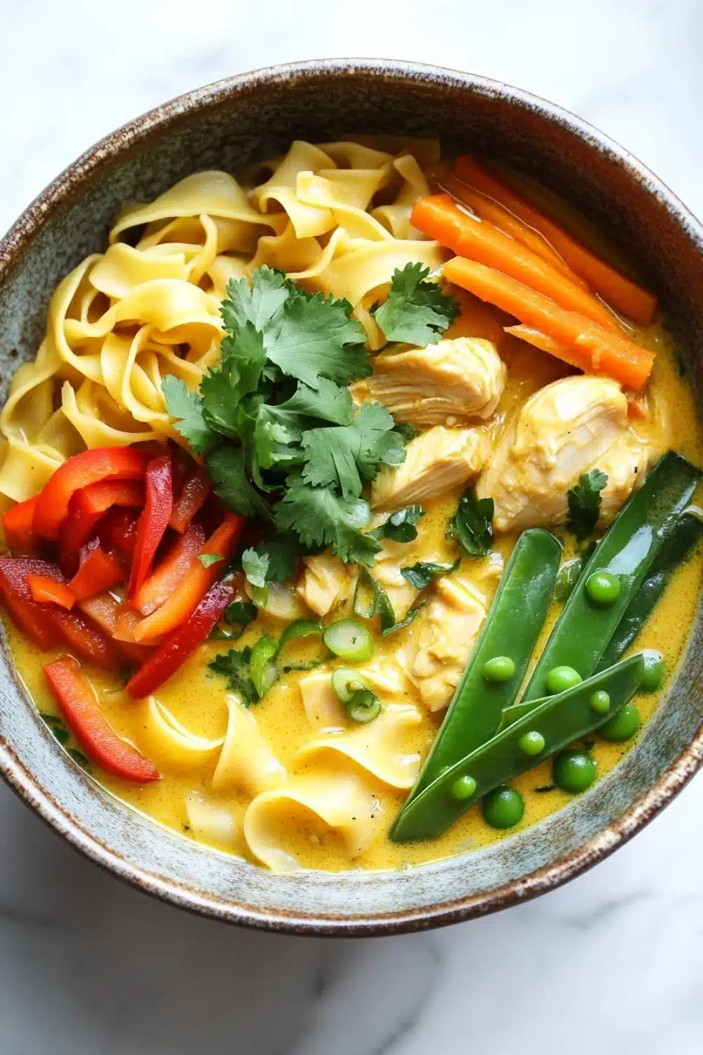 Yellow curry chicken noodle soup - the image is a close-up of a bowl of soup. the soup appears to be a creamy yellow color with chunks of chicken and vegetables mixed in. the bowl is made of ceramic and has a brown rim. on top of the soup, there are sliced carrots, red bell peppers, and green peas. there is also a sprig of cilantro on top. the noodles are thin and yellow, and the soup is garnished with fresh cilantro leaves. the background is a white marble countertop.