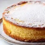Yellow lemon cake design - the image is a close-up of a round cake on a white plate. the cake appears to be a yellowish-orange color and has a smooth texture. it is dusted with a generous amount of powdered sugar, giving it a shiny appearance. the plate is sitting on a marble countertop, and the background is blurred, making the cake the focal point of the image.