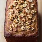 Zucchini bread almond flour - the image is a close-up of a loaf of bread on a wooden surface. the bread appears to be freshly baked and has a golden brown crust on top. it is topped with chopped walnuts, giving it a crunchy texture. the walnuts are arranged in a scattered manner, with some overlapping each other. the loaf is slightly tilted to the side, and the background is blurred, making the bread the focal point of the image.
