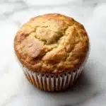 Zucchini lemon muffin - the image is a close-up of a freshly baked muffin on a white marble surface. the muffin is round and golden brown in color, with a crumbly texture on top. it appears to be freshly baked and has a soft, fluffy interior. the edges of the muffin are lined with white paper liners. the background is blurred, making the muffins the focal point of the image.