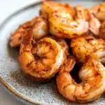 Air fryer carribean jerk shrimp - the image shows a plate of cooked shrimp on a white tablecloth. the shrimp are arranged in a circular pattern on the plate, with some overlapping each other. they are golden brown and appear to be seasoned with herbs and spices. the plate is made of a light-colored ceramic or porcelain material with a speckled texture. the background is blurred, making the shrimp the focal point of the image.