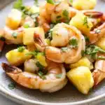 Air fryer hawaiian shrimp - the image shows a plate of cooked shrimp and pineapple salad. the shrimp are cooked medium-sized and have a pinkish-orange color. they are arranged in a circular pattern on the plate, with some pieces of pineapple scattered around them. the pineapple is cut into small cubes and is garnished with a sprig of fresh parsley. the dish is served on a white marble countertop.