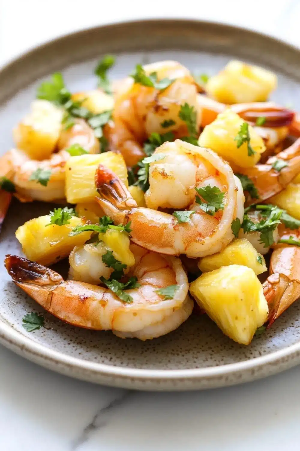 Air fryer hawaiian shrimp - the image shows a plate of cooked shrimp and pineapple salad. the shrimp are cooked medium-sized and have a pinkish-orange color. they are arranged in a circular pattern on the plate, with some pieces of pineapple scattered around them. the pineapple is cut into small cubes and is garnished with a sprig of fresh parsley. the dish is served on a white marble countertop.