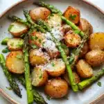 Air fryer potato and asparagus - the image is a close-up of a plate of roasted potatoes and asparagus. the potatoes are golden brown and appear to be seasoned with herbs and grated parmesan cheese. the asparaglips are arranged in a circular pattern on the plate, with some of them overlapping each other. the plate is white with a light blue rim and is placed on a white marble countertop. there is a fork and knife on the side of the plate. the overall presentation of the dish is elegant and appetizing.