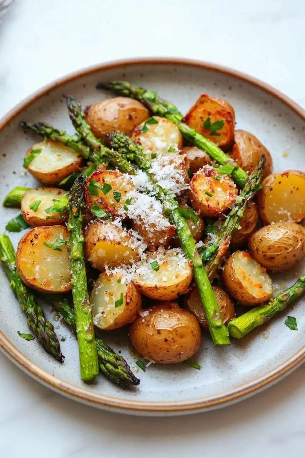Air fryer potato and asparagus - the image is a close-up of a plate of roasted potatoes and asparagus. the potatoes are golden brown and appear to be seasoned with herbs and grated parmesan cheese. the asparaglips are arranged in a circular pattern on the plate, with some of them overlapping each other. the plate is white with a light blue rim and is placed on a white marble countertop. there is a fork and knife on the side of the plate. the overall presentation of the dish is elegant and appetizing.