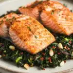 Air fryer salmon and swiss chard - the image shows two pieces of salmon on a plate. the salmon is cooked to a golden brown color and appears to be seasoned with herbs and spices. the plate is white and has a textured surface. on top of the salmon, there is a bed of greens, including collard greens, red onions, and pine nuts. the greens are mixed with the salmon and the pine nuts, creating a colorful and appetizing dish. the background is blurred, making the salmon the focal point of the image.