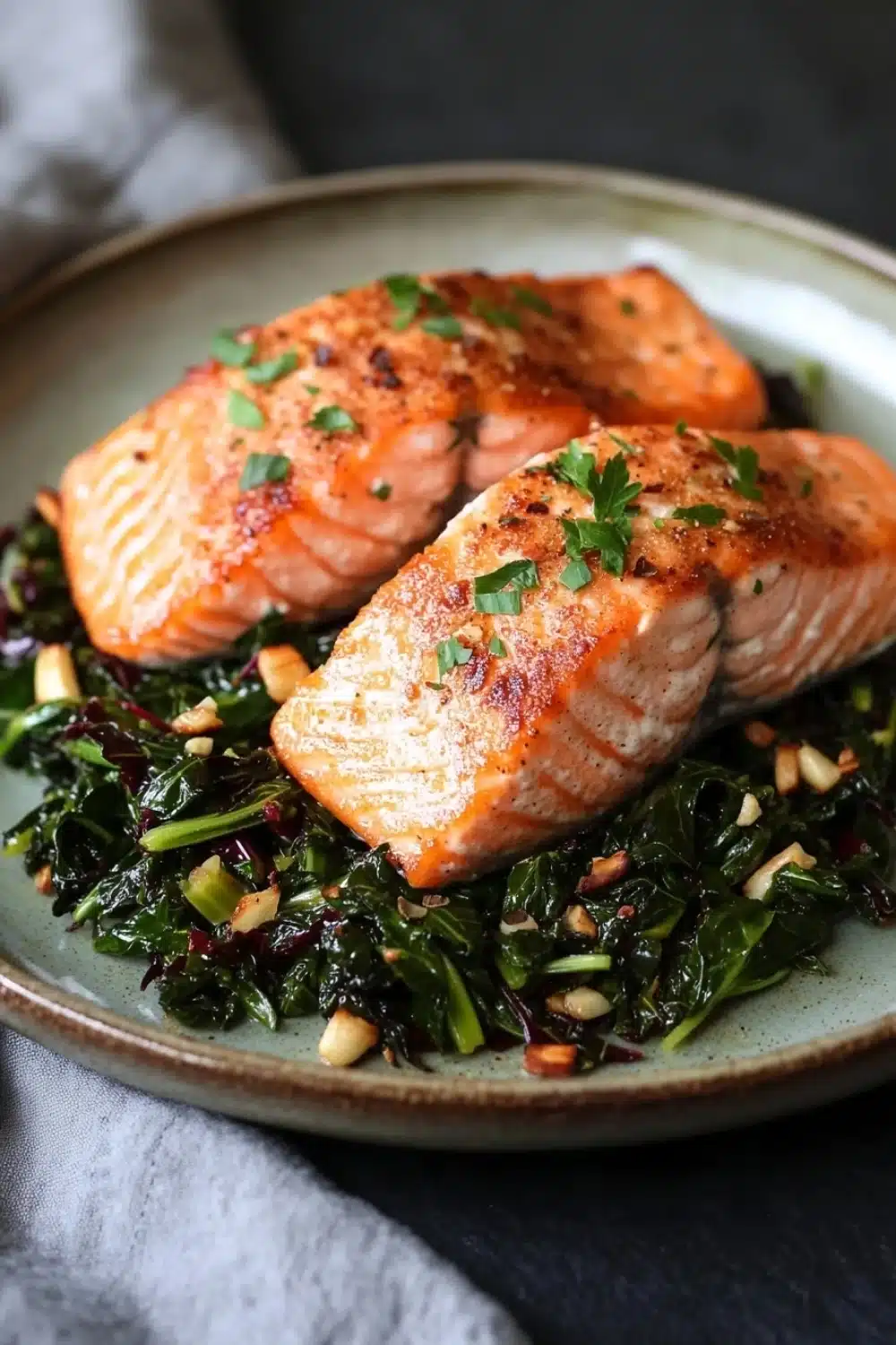 Air fryer salmon and swiss chard - the image shows two pieces of salmon on a plate. the salmon is cooked to a golden brown color and appears to be seasoned with herbs and spices. the plate is light blue and has a textured surface. on top of the salmon, there is a bed of greens, including collard greens, red cabbage, and pine nuts. the greens are garnished with chopped parsley. the background is dark and out of focus, making the salmon and greens the focal point of the image.