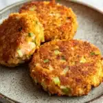 Air fryer salmon cakes - the image shows three round, golden-brown, fried food items on a blue and white speckled plate. the food appears to be a type of crab cake, with a crispy exterior and a soft, fluffy interior. the top of the food is garnished with chopped green onions, adding a pop of color to the dish. the plate is sitting on a white marble countertop.