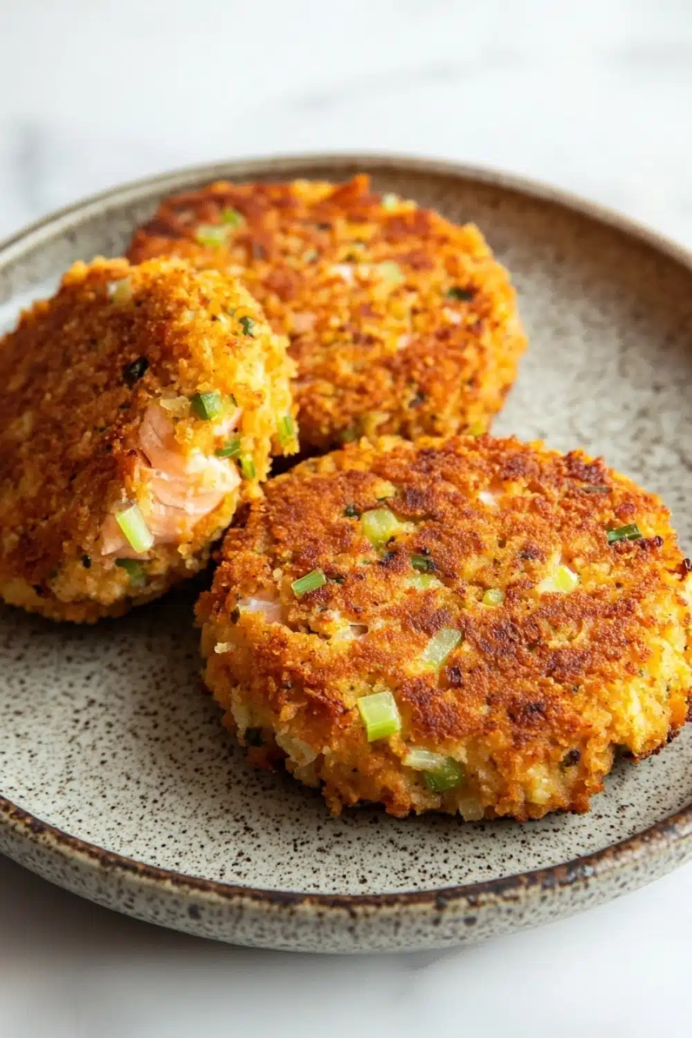 Air fryer salmon cakes - the image shows three round, golden-brown, fried food items on a blue and white speckled plate. the food appears to be a type of crab cake, with a crispy exterior and a soft, fluffy interior. the top of the food is garnished with chopped green onions, adding a pop of color to the dish. the plate is sitting on a white marble countertop.