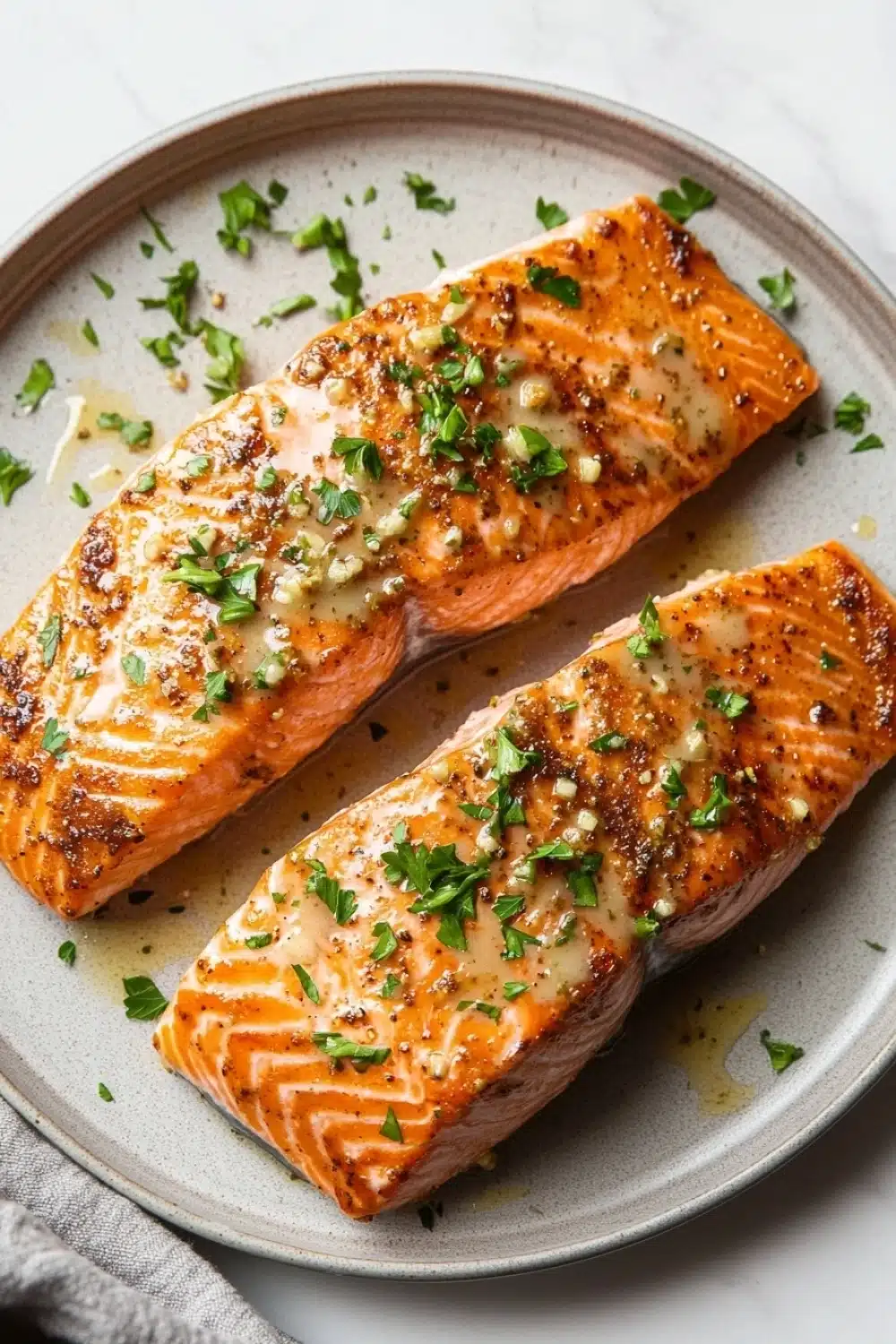 Air fryer salmon garlic butter - the image shows two pieces of salmon on a gray plate. the salmon is cooked to a golden brown color and appears to be seasoned with herbs and spices. the plate is garnished with chopped parsley and grated parmesan cheese. the background is a white marble countertop with a gray napkin on the side.