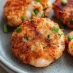 Air fryer shrimp patties - the image shows a plate of four crab cakes. the crab cakes are golden brown and appear to be crispy on the outside. they are garnished with chopped green onions. the plate is white with a speckled pattern and is sitting on a wooden table. the background is blurred, but it appears to be a kitchen countertop.