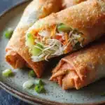 Air fryer vegetable spring rolls - the image shows a plate with three spring rolls on it. the spring rolls are golden brown and appear to be crispy on the outside. they are filled with a variety of vegetables, including carrots, green onions, and shredded cheese. the plate is garnished with chopped green onions. the background is a dark blue tablecloth.