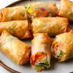Air fryer vegetable spring rolls - the image shows a plate of freshly baked spring rolls. the rolls are golden brown and appear to be crispy on the outside. they are filled with a variety of vegetables, including carrots, celery, and onions, and are arranged in a circular pattern on the plate. the plate is white and has a textured surface. the background is blurred, but it appears to be a kitchen countertop.