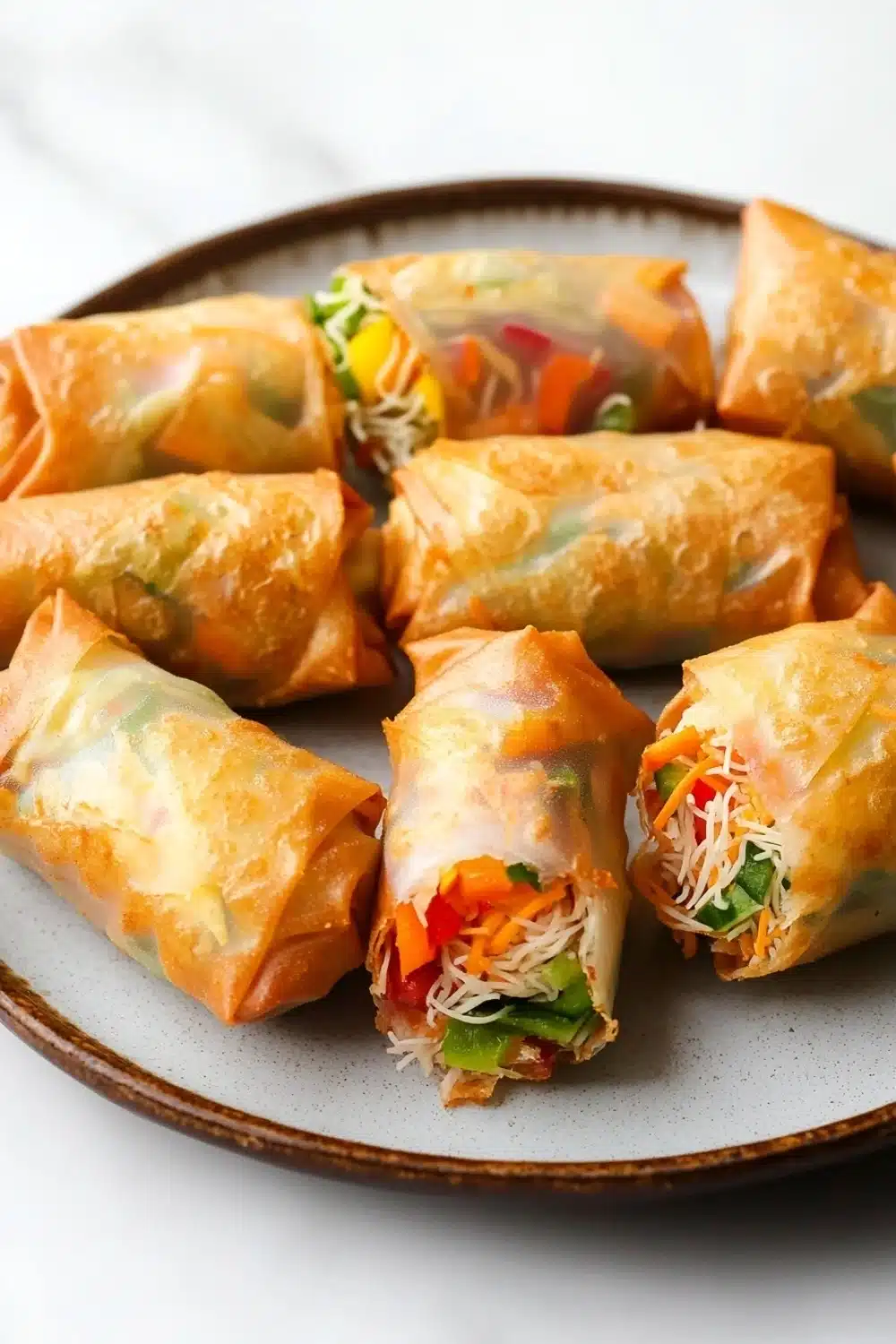 Air fryer vegetable spring rolls - the image shows a plate of freshly baked spring rolls. the rolls are golden brown and appear to be crispy on the outside. they are filled with a variety of vegetables, including carrots, celery, and onions, and are arranged in a circular pattern on the plate. the plate is white and has a textured surface. the background is blurred, but it appears to be a kitchen countertop.