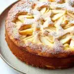 Apple cake korean - the image is of a round cake on a white plate. the cake appears to be freshly baked and has a golden brown crust. on top of the cake, there is a lattice-like pattern made up of sliced apples arranged in a symmetrical manner. the apples are dusted with a light dusting of powdered sugar. the plate is sitting on a marble countertop.