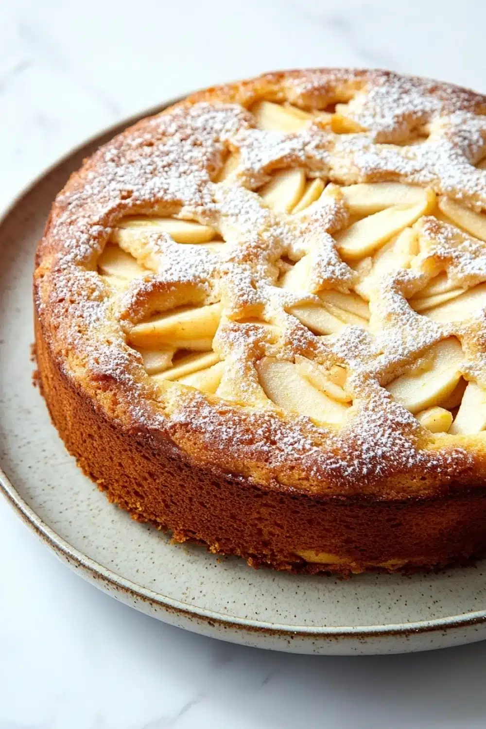 Apple cake korean - the image is of a round cake on a white plate. the cake appears to be freshly baked and has a golden brown crust. on top of the cake, there is a lattice-like pattern made up of sliced apples arranged in a symmetrical manner. the apples are dusted with a light dusting of powdered sugar. the plate is sitting on a marble countertop.
