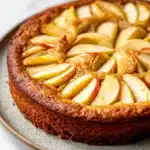 Apple cake korean - the image is of a round cake on a white plate. the cake appears to be a tart with a golden brown crust and a layer of caramelized apples on top. the apples are arranged in a circular pattern on the top of the cake, with some overlapping each other. the caramel is drizzled over the apples, giving them a glossy sheen. the plate is sitting on a marble countertop.