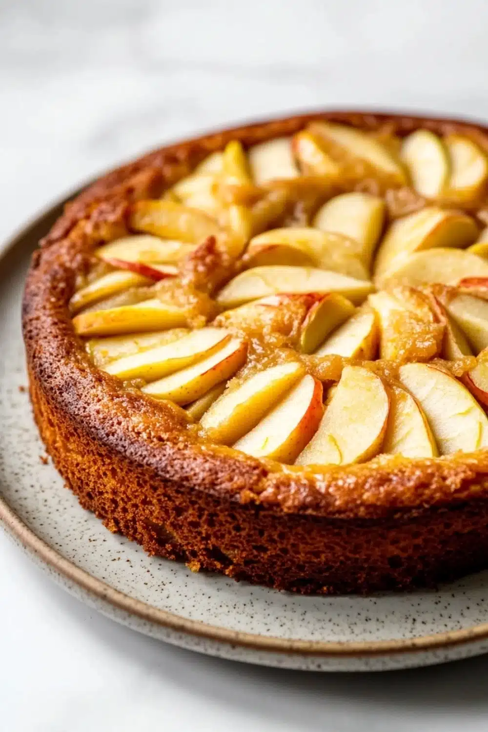 Apple cake korean - the image is of a round cake on a white plate. the cake appears to be a tart with a golden brown crust and a layer of caramelized apples on top. the apples are arranged in a circular pattern on the top of the cake, with some overlapping each other. the caramel is drizzled over the apples, giving them a glossy sheen. the plate is sitting on a marble countertop.