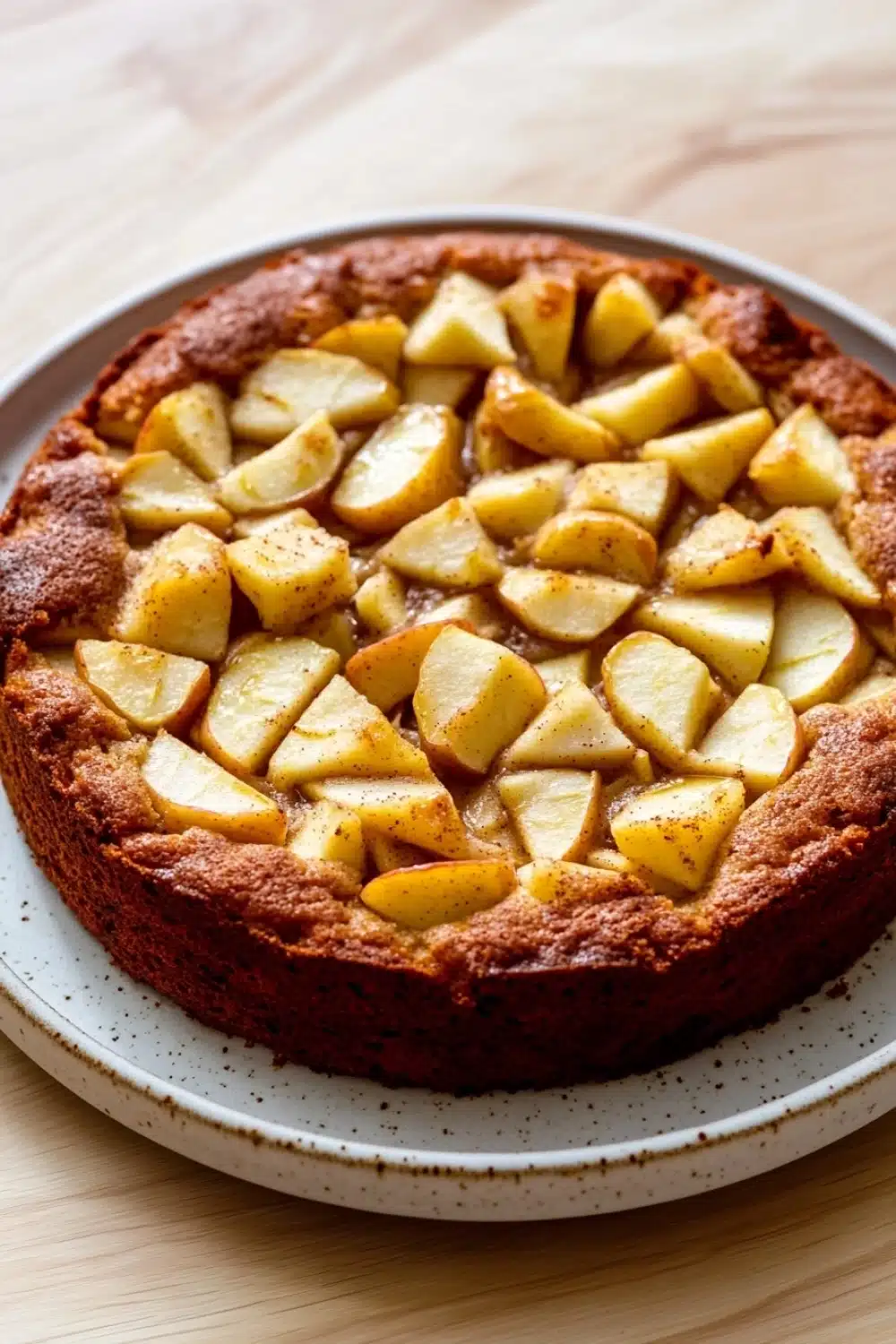 Apple cake sugar free - the image is of a round cake on a white plate. the cake appears to be freshly baked and has a golden brown crust on top. on top of the cake, there are small cubes of sliced apples arranged in a circular pattern. the apples are cut into small cubes and are evenly spaced along the edges of the crust. the plate is sitting on a wooden table.