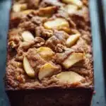 Apple cinnamon muffin loaf - the image shows a rectangular loaf of bread on a black baking tray. the bread appears to be freshly baked and has a golden brown color. on top of the bread, there are sliced apples arranged in a crumbly pattern, with some of the apples overlapping each other. the apples are cut into small cubes and are scattered throughout the bread. the tray is lined with parchment paper, and the background is blurred, making the bread the focal point of the image.