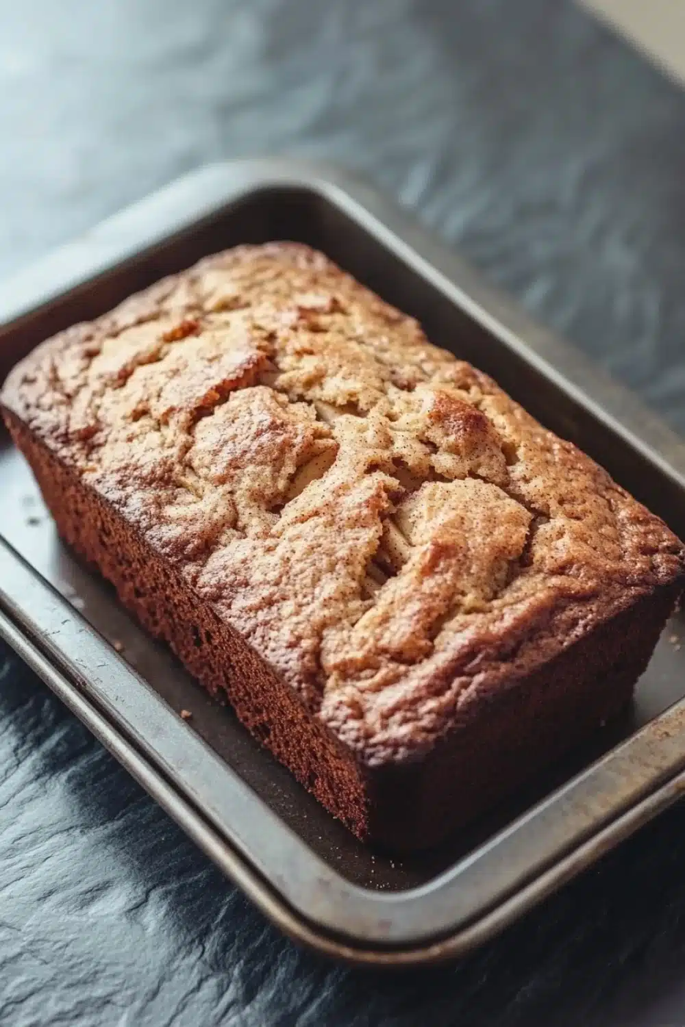 Apple cinnamon muffin loaf - the image shows a freshly baked loaf of bread on a metal baking tray. the bread appears to be golden brown in color and has a crumbly texture on top. the tray is placed on a dark grey countertop with a textured surface. the background is blurred, but it seems to be a kitchen countertop.