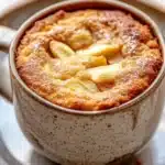 Apple mug cake - the image is a close-up of a dessert in a ceramic mug. the mug is sitting on a white plate with a gold rim. the dessert appears to be a round, golden-brown cake with a crumbly texture. on top of the cake, there are sliced apples arranged in a circular pattern. the apples are covered in a light dusting of powdered sugar. the background is blurred, but it seems to be an indoor setting with a wooden table.