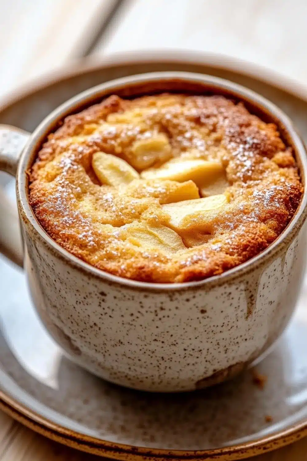 Apple mug cake - the image is a close-up of a dessert in a ceramic mug. the mug is sitting on a white plate with a gold rim. the dessert appears to be a round, golden-brown cake with a crumbly texture. on top of the cake, there are sliced apples arranged in a circular pattern. the apples are covered in a light dusting of powdered sugar. the background is blurred, but it seems to be an indoor setting with a wooden table.