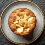 Apple mug cake - the image is a close-up of a round cake on a plate. the cake appears to be freshly baked and has a golden brown crust on top. it is cut into small cubes and is arranged in a circular pattern on the plate, with some of the cubes overlapping each other. the plate is made of a light-colored ceramic with a speckled pattern around the edges. the background is a dark grey textured surface. the overall mood of the image is rustic and cozy.