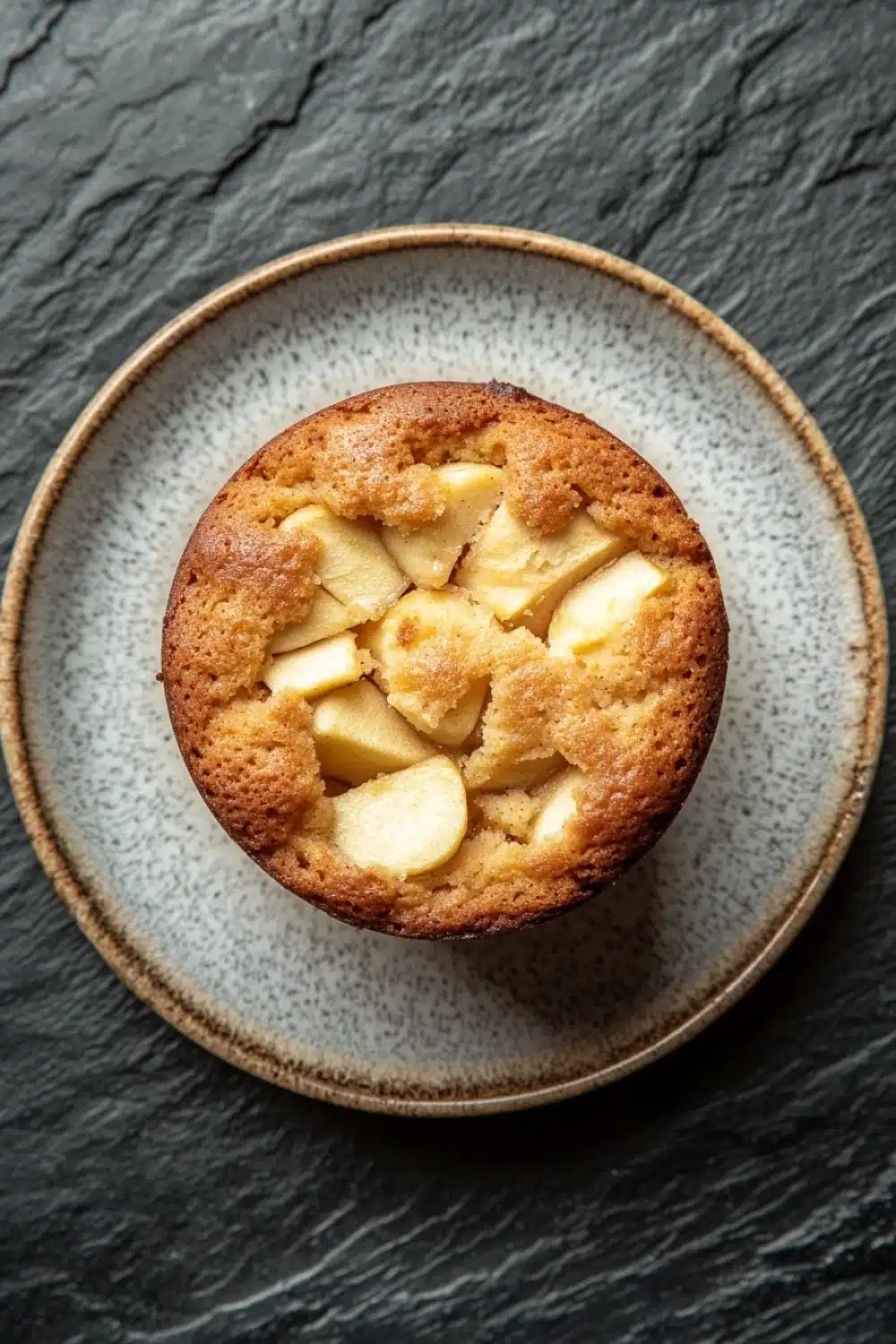 Apple mug cake - the image is a close-up of a round cake on a plate. the cake appears to be freshly baked and has a golden brown crust on top. it is cut into small cubes and is arranged in a circular pattern on the plate, with some of the cubes overlapping each other. the plate is made of a light-colored ceramic with a speckled pattern around the edges. the background is a dark grey textured surface. the overall mood of the image is rustic and cozy.
