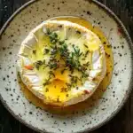 Baked brie with honey and thyme - the image is a top view of a dish on a wooden table. the dish appears to be a poached egg with a golden-brown yolk and a sprig of fresh thyme on top. the plate is round and has a speckled texture. the background is dark and out of focus, making the dish stand out. the overall mood of the image is rustic and appetizing.