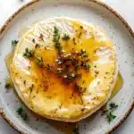 Baked brie with honey and thyme - the image is a close-up of a plate of food. the plate is white with a brown rim and has a speckled pattern. on top of the plate, there is a poached egg with a golden-brown yolk and a drizzle of yellow sauce. the egg is garnished with a sprig of fresh thyme. the background is a white marble countertop with a few sprigs of green herbs scattered around. the overall aesthetic of the image is rustic and appetizing.