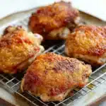 Baked chicken thighs on wire rack - the image shows four pieces of chicken on a wire cooling rack. the chicken appears to be cooked and has a golden brown color with sesame seeds sprinkled on top. the rack is placed on a white countertop. the background is blurred, so the focus is on the chicken.