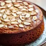 Banana cake almond - the image is of a round cake on a blue plate. the cake appears to be a chocolate cake with a golden brown crust and is topped with sliced almonds. the almonds are arranged in a circular pattern on top of the cake, creating a textured surface. the plate is sitting on a wooden table with a blurred background.