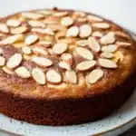 Banana cake almond - the image is of a round cake on a white plate. the cake appears to be a chocolate cake with a golden brown crust and is topped with sliced almonds. the almonds are arranged in a circular pattern on top of the cake, creating a lattice-like design. the plate is sitting on a marble countertop. the background is blurred, making the cake the focal point of the image.