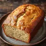 Banana cake loaf - the image shows a freshly baked loaf of bread on a gray plate. the bread appears to be golden brown in color and has a slightly crumbly texture. it has a soft, fluffy interior with visible chunks of butter and nuts scattered throughout. the plate is placed on a wooden table with a rustic, weathered look. the background is blurred, making the bread the focal point of the image.