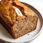 Banana cake loaf - the image shows a freshly baked loaf of banana bread on a white marble countertop. the bread is golden brown in color and has a slightly crumbly texture. it has a slice taken out of it, revealing the soft, fluffy interior. a single banana peel is visible on top of the bread, adding a pop of color to the dish. the plate it is sitting on is round and has an speckled pattern. the background is blurred, making the bread the focal point of the image.