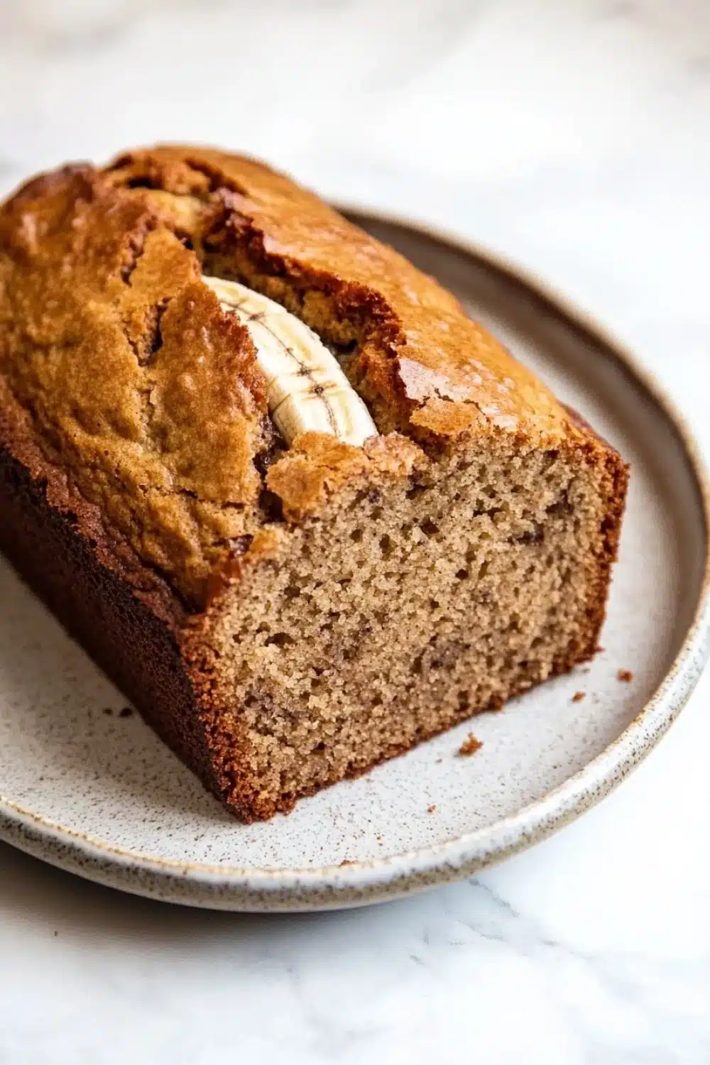 Banana cake loaf - the image shows a freshly baked loaf of banana bread on a white marble countertop. the bread is golden brown in color and has a slightly crumbly texture. it has a slice taken out of it, revealing the soft, fluffy interior. a single banana peel is visible on top of the bread, adding a pop of color to the dish. the plate it is sitting on is round and has an speckled pattern. the background is blurred, making the bread the focal point of the image.