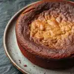Banana cake no butter - the image is a close-up of a round cake on a white plate with a blue rim. the cake appears to be a chocolate cake with a golden brown crust on top. the top of the cake is covered in a light dusting of powdered sugar, giving it a shiny appearance. the plate is sitting on a dark grey textured surface. the background is blurred, making the cake the focal point of the image.