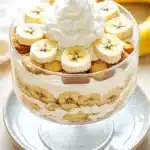 Banana cake trifle - the image is of a dessert in a glass trifle dish. the trifle is topped with a generous dollop of whipped cream and sliced bananas. the bananas are arranged in a circular pattern on top of the trifle, with some of them overlapping each other. there are also some crumbs scattered throughout the dessert. the dish is sitting on a white plate with a blue rim. in the background, there is a bunch of bananas and a white napkin. the overall color scheme of the image is warm and inviting.