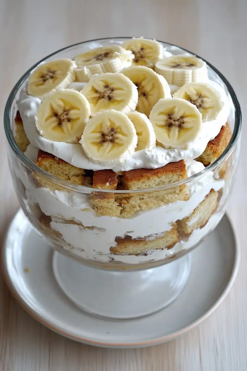 Banana cake trifle - the image is of a dessert in a glass cake stand. the cake stand is white with a gold rim and is placed on a wooden table. the top of the cake stand has a layer of whipped cream and sliced bananas arranged in a circular pattern. the bananas are yellow and appear to be ripe and juicy. there are also a few crumbs scattered around the cake, indicating that it has been freshly baked. the overall appearance of the dessert is fresh and appetizing.