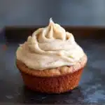 Banana muffin frosting - the image is a close-up of a cupcake on a black baking tray. the cupcake has a golden brown crust and is topped with a generous swirl of white frosting. the frosting appears to be smooth and creamy, with small black speckles scattered throughout. the background is blurred, making the cupcake the focal point of the image.