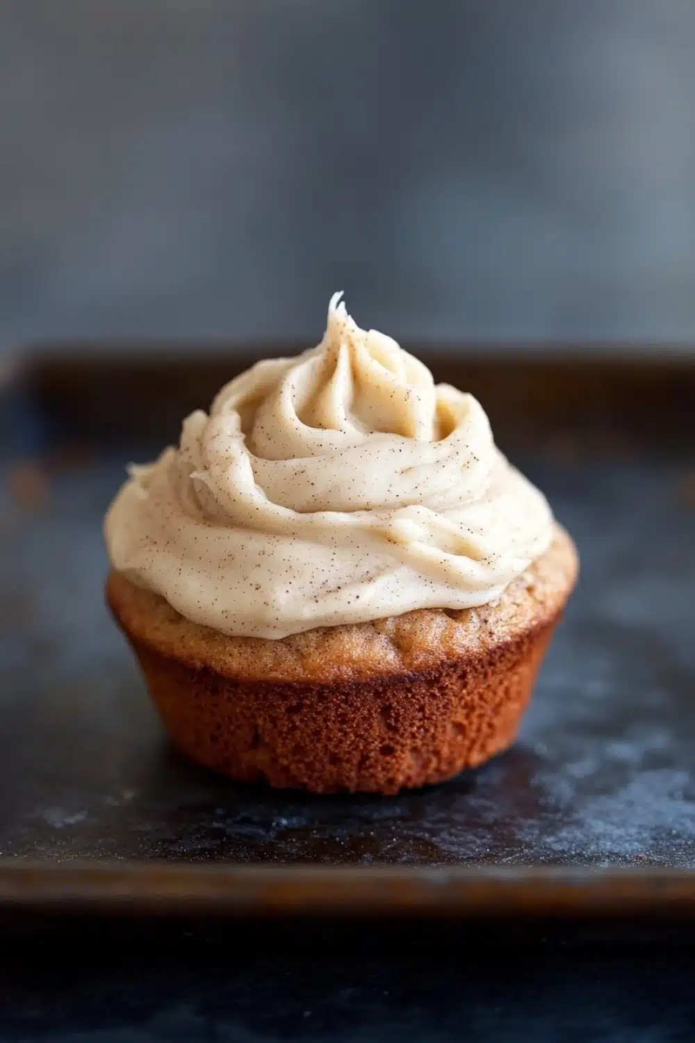 Banana muffin frosting - the image is a close-up of a cupcake on a black baking tray. the cupcake has a golden brown crust and is topped with a generous swirl of white frosting. the frosting appears to be smooth and creamy, with small black speckles scattered throughout. the background is blurred, making the cupcake the focal point of the image.