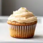 Banana muffin frosting - the image is of a cupcake on a white plate. the cupcake has a golden brown crust and is topped with a generous layer of creamy white frosting. the frosting is swirled in a swirl pattern and has small black speckles scattered throughout. the plate is sitting on a marble countertop. the background is blurred, but it appears to be a kitchen countertop with a gray wall.