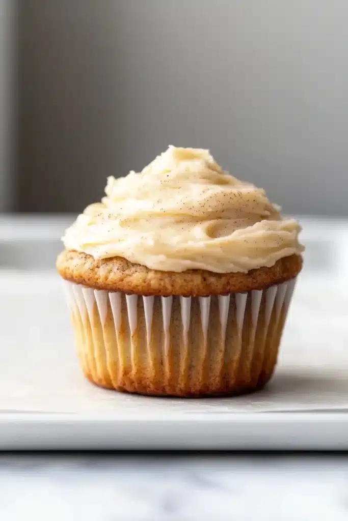Banana muffin frosting - the image is of a cupcake on a white plate. the cupcake has a golden brown crust and is topped with a generous layer of creamy white frosting. the frosting is swirled in a swirl pattern and has small black speckles scattered throughout. the plate is sitting on a marble countertop. the background is blurred, but it appears to be a kitchen countertop with a gray wall.