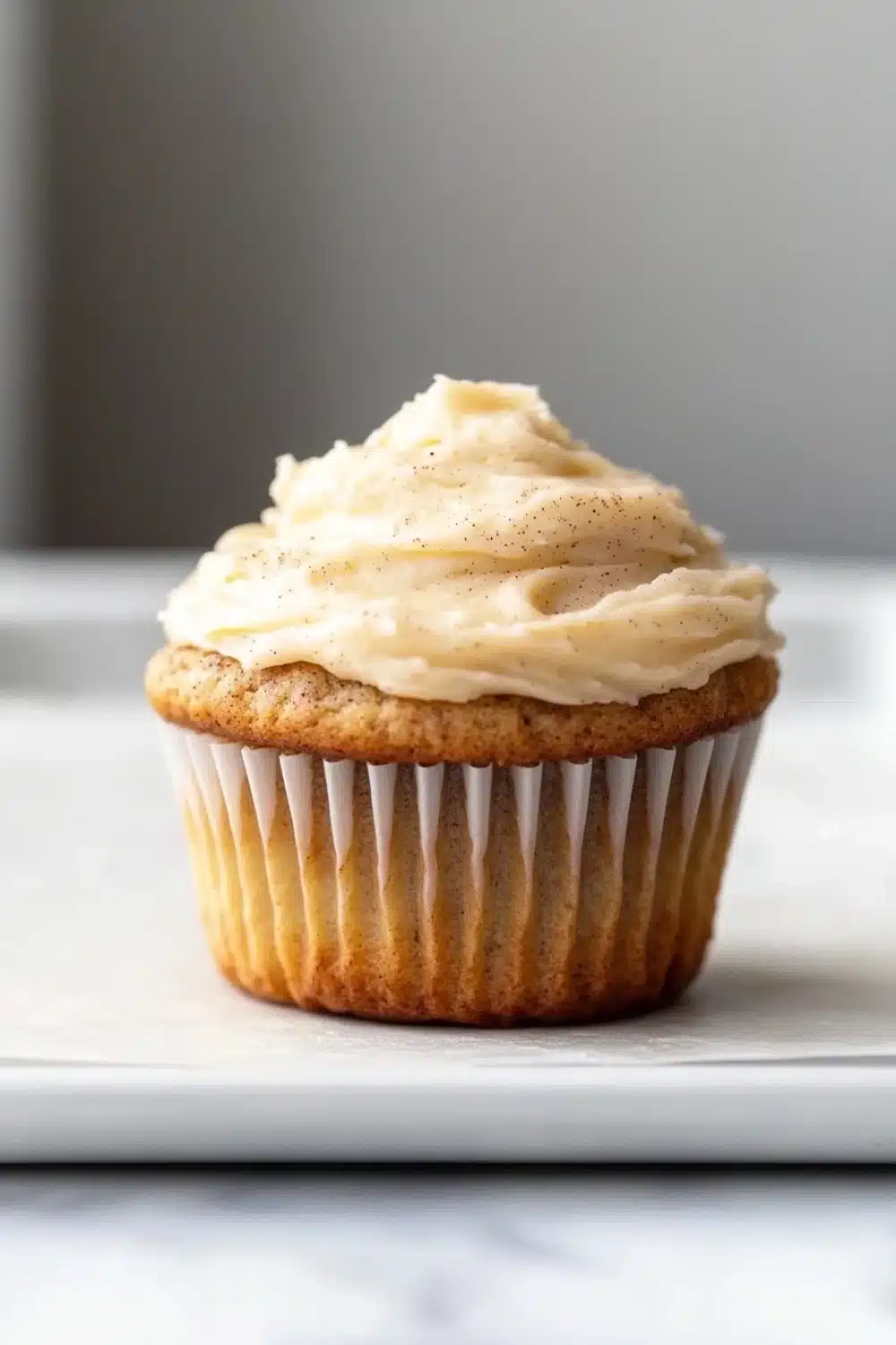 Banana muffin frosting - the image is of a cupcake on a white plate. the cupcake has a golden brown crust and is topped with a generous layer of creamy white frosting. the frosting is swirled in a swirl pattern and has small black speckles scattered throughout. the plate is sitting on a marble countertop. the background is blurred, but it appears to be a kitchen countertop with a gray wall.