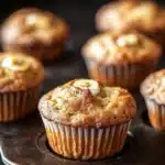 Banana muffin sour cream - the image shows a group of freshly baked muffins on a black baking tray. the muffins are golden brown in color and have a crumbly texture. they are topped with a generous amount of butter, which is visible on top of each muffin. the tray is lined with parchment paper and is placed on a wooden surface. the background is blurred, but it appears to be a kitchen countertop.