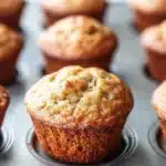 Banana muffin sour cream - the image shows a muffin tin filled with freshly baked muffins. the muffins are golden brown in color and have a crumbly texture on top. they are arranged in a single layer in the tin, with some overlapping each other. the tin is placed on a white marble countertop. the background is blurred, but it appears to be a kitchen countertop with more muffins visible.