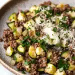 Beef and zucchini skillet - the image is a close-up of a plate of food. the plate is made of a light-colored ceramic bowl with a brown rim. the food appears to be a dish made with zucchini, ground beef, and parmesan cheese. the zucchinis are cut into small cubes and are arranged in a pile on top of the ground beef. the ground beef is a dark brown color and is scattered throughout the dish. there are also some chopped parsley scattered throughout, adding a pop of green color to the dish and giving it a fresh and appetizing appearance. the background is a white marble countertop.